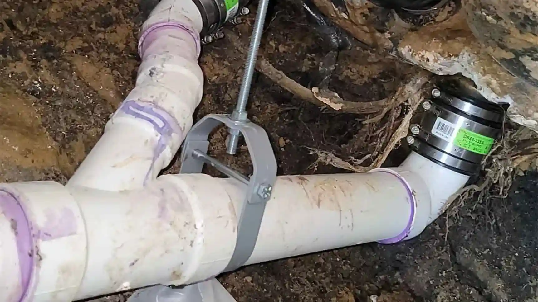 A new, white PVC pipe installed inreplacement of cast iron pipe in excavated ground, connected with black couplings and supported by a metal bracket.