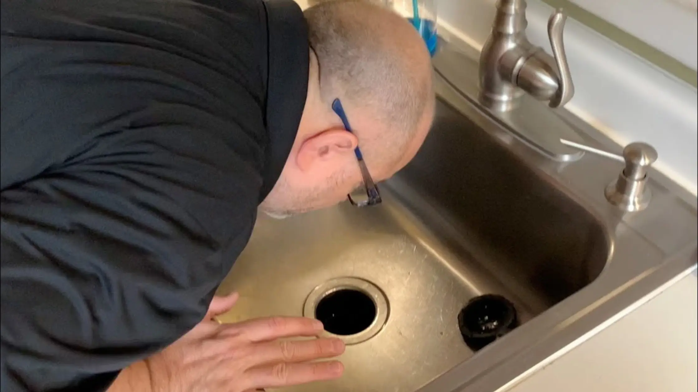  A man in a black shirt and blue glasses leans over a kitchen sink to inspect the dark drain smell.