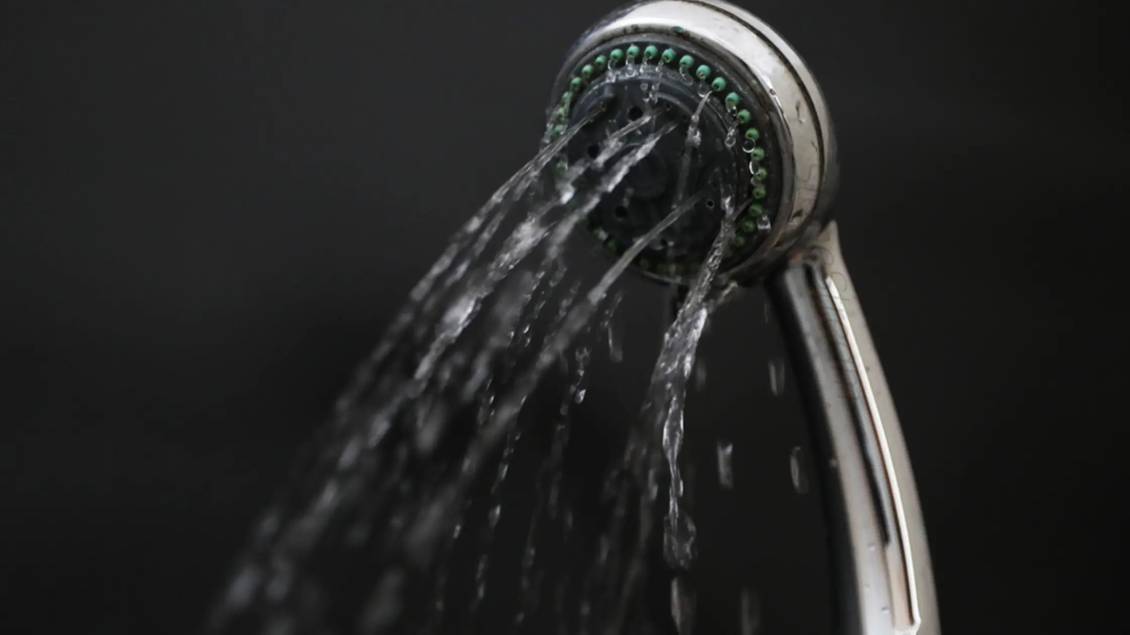 A silver shower head sprays a weak stream of water against a dark background.