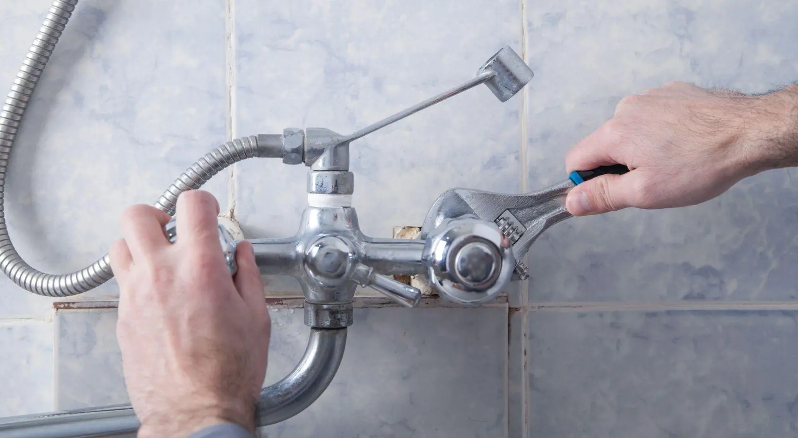  A pair of hands uses a wrench to tighten or loosen a fitting on a chrome shower faucet, suggesting a plumbing repair.